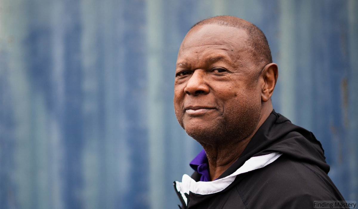 Coach George Mumford in a black jacket stands against a textured blue background, conveying a sense of confidence