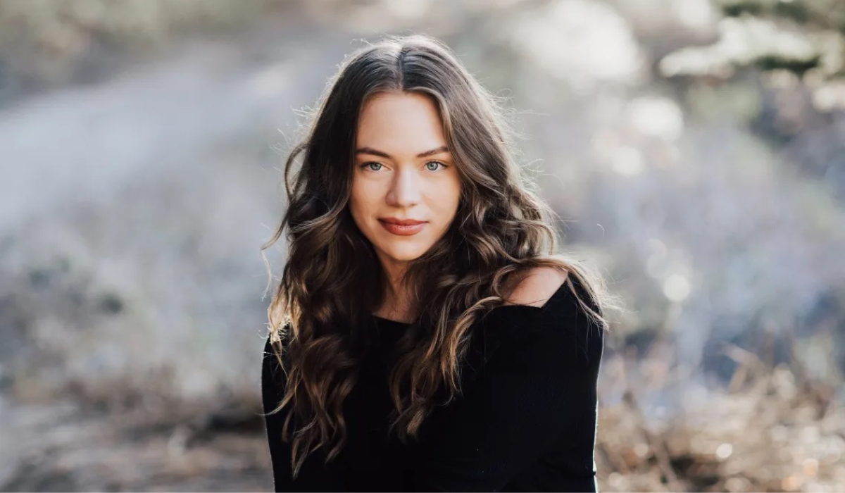 Brianna Weist with long, wavy hair wears a black off-shoulder top, seated outdoors in a softly blurred natural background