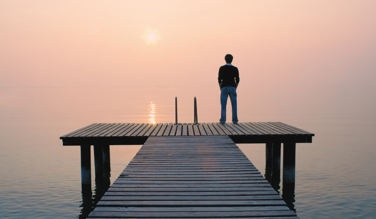 Man stares off a dock overlooking a pink haze covering the lake and sky.