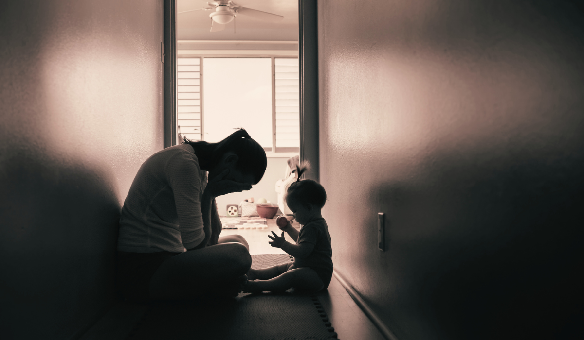 A frustrated mother hangs her head in her hands as a todler plays next to her.